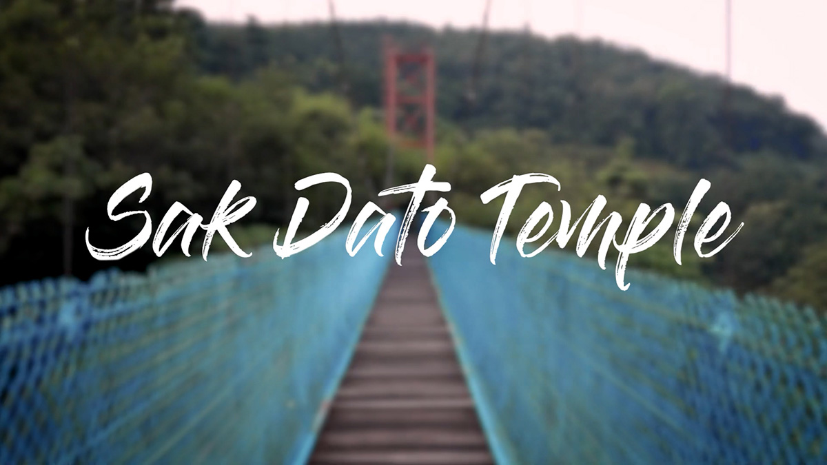 A text that reads 'Sak Dato Temple' on top of a blurry background of a bridge in nature.
